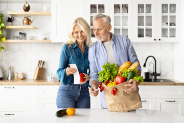 Smiling Senior Couple In Kitchen Checking Bills After Food Shopping