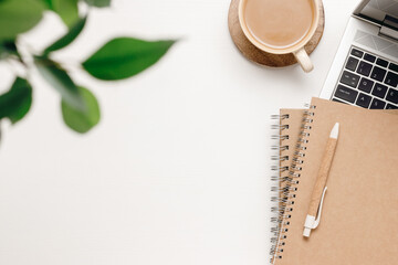 Workspace with notepad,coffee, laptop, office supplies and green plant leaves on white wooden table, copy space. Home office, creative workplace, aesthetic style, top view.