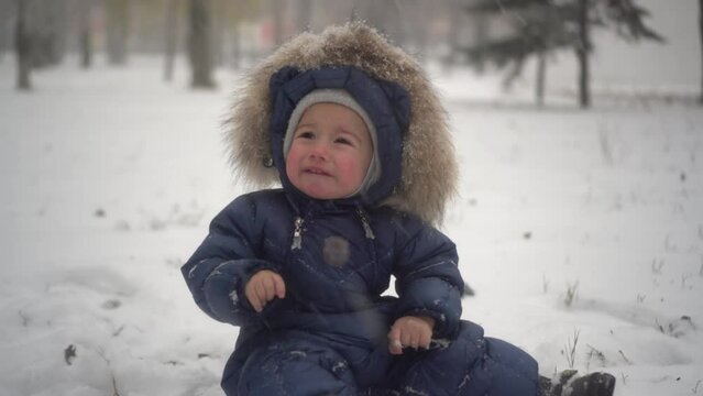 A small child sits in the snow in the park and cries from the cold. The boy in the down overalls had cold hands.