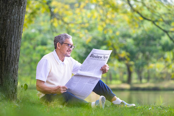 Senior Asian man reading a business newspaper while sitting under the tree by the lake at the public park for recreation, leisure and relaxation in nature