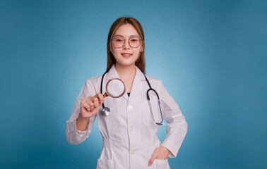 Young asian doctor put hand in pocket of medical uniform lab coat on blue background. Smiling female doctor holding magnifier to check for disease. Doctor examining the health of the patient.