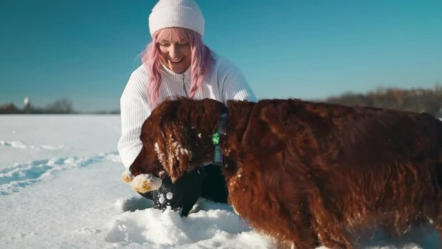 Happy beautiful young woman blowing snowflakes from her hands to her dog irish setter enjoying winter snow outdoors activity in winter day holiday. People and animals together having fun and smiling