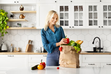 Cheerful senior woman unpacking paper bag full of organic vegetables in kitchen