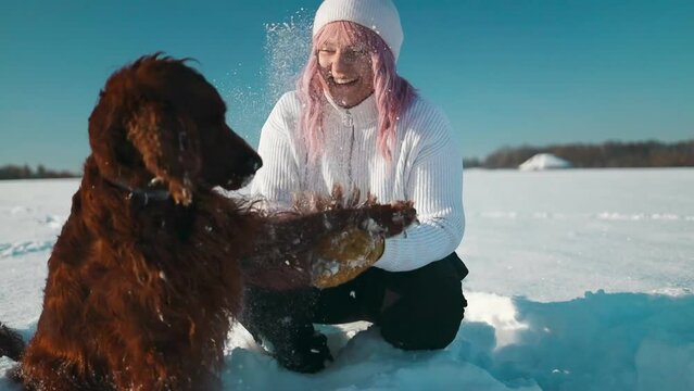 Happy beautiful young woman blowing snowflakes from her hands to her dog irish setter enjoying winter snow outdoors activity in winter day holiday. People and animals together having fun and smiling