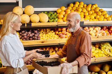 bearded cheerful seller helping his female customer to choose blackberries, smiling at each other