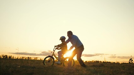 Attentive father silhouette teaches son to ride bike in field at sunset dusk - obrazy, fototapety, plakaty