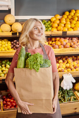 mature cheerful saleswoman posing with paper bag full of greens and looking away at grocery shop