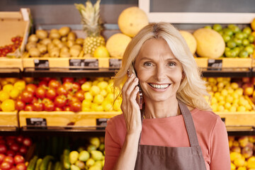 joyful mature blonde saleswoman talking by mobile phone and smiling at camera at grocery store