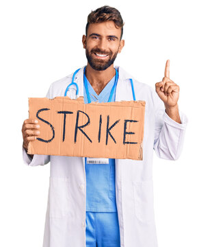 Young hispanic man wearing doctor stethoscope holding strike banner surprised with an idea or question pointing finger with happy face, number one
