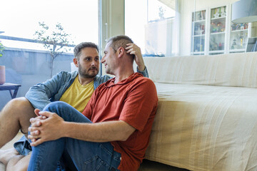 adult homosexual couple caressing each other sitting on the floor of their living room looking at...
