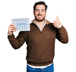 Young hispanic man holding travel calendar smiling happy and positive, thumb up doing excellent and approval sign