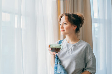 Free Time To Relax. Young woman looking out the window while overlooking the city at cozy home holding cup of hot tea drink. Happy calm female taking break for mental health wellbeing. No stress.