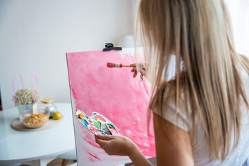 Young woman artist with palette and brush painting abstract pink picture on canvas at home. Back view. Art and creativity concept