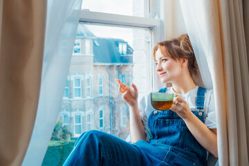Free Time To Relax. Young woman looks out the window overlooking the city, sits on the windowsill...