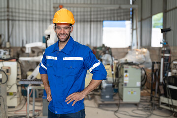 Portrait of male engineer in uniform smiling and arms akimbo at industrial factory. Technician man standing wearing yellow helmet safety in manufacturing workshop.