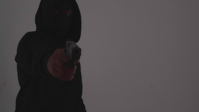 Mysterious man wearing black hoodie holding a pistol wearing a cloth mask covering his face completely in front of the door, Silhouette and dark concept image,threat of firearms,attack or defense.