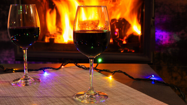 Two red wine glasses and lights decoration on burning fireplace background. Wine by the fireplace.