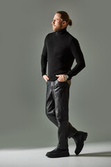 handsome bearded man in black turtleneck sweater and leather pants posing on grey background