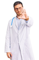 Handsome young man with bear wearing doctor uniform doing stop sing with palm of the hand. warning expression with negative and serious gesture on the face.