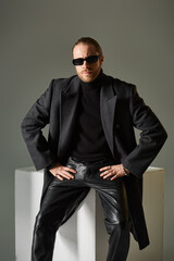 good looking bearded man in stylish sunglasses and coat sitting on white cube on grey backdrop