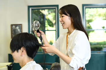 男性客の髪をドライヤーで乾かすアジア人の女性美容師 - obrazy, fototapety, plakaty