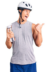 Young handsome man wearing bike helmet holding bottle of water pointing thumb up to the side smiling happy with open mouth