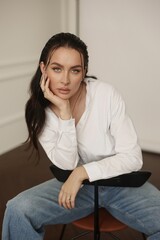 Fototapeta premium Beautiful brunet woman is captured in a photo studio, dressed in blue jeans and a white blouse. She exudes elegance and grace as she sits gracefully on a chair