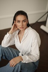 In a photo studio, a young woman with dark brown hair exudes charm in her blue denim jeans and a pristine white blouse as she confidently poses on the floor.