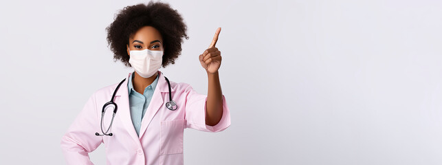 African American woman doctor in a mask and pink robe points her finger to the side on a white background. Free space for product placement or advertising text.