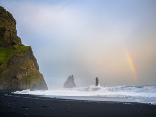 Strong surf at a Reynisfjara black sand beach. Rainbow showing on right side. Reynisdrangar Sea Stacks getting hit by waves. South Coast of Iceland.