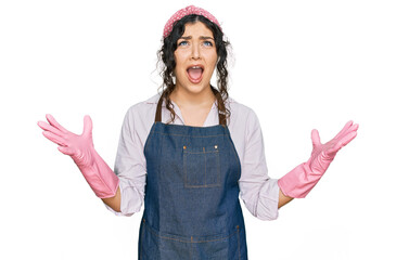 Young hispanic girl wearing cleaner apron and gloves crazy and mad shouting and yelling with...