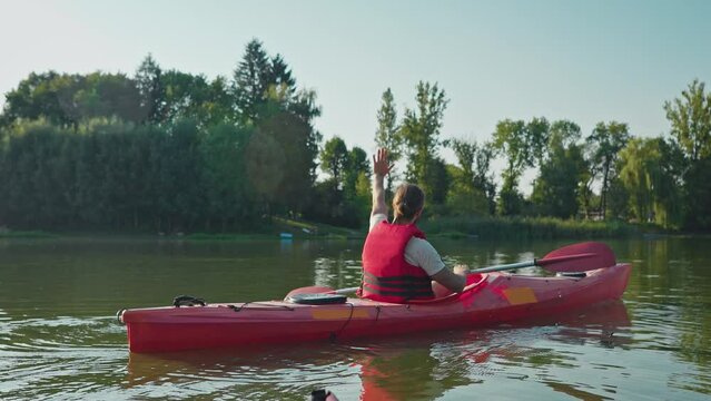 Side view of young man with red safety vest traveling on boat among large lake or river. Male holding oar and paddling in kayak. Waving hand to people on pond bank. Concept of relaxation.