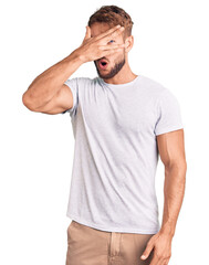 Young caucasian man wearing casual white tshirt peeking in shock covering face and eyes with hand, looking through fingers with embarrassed expression.