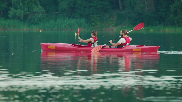 Side view of couple rowing on double kayak. Bearded man relaxing with his woman outside in boat. Swimming on kayak in middle of reservoir. People having summer holiday. Leisure concept.