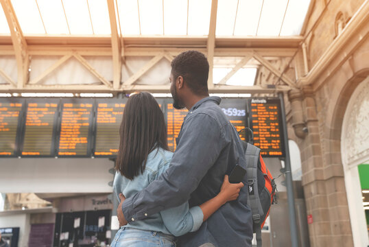Happy man and woman in love waiting for a train and cuddle at a railway station in front of the timetable toned image