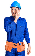 Young handsome man wearing worker uniform and hardhat hand on mouth telling secret rumor, whispering malicious talk conversation