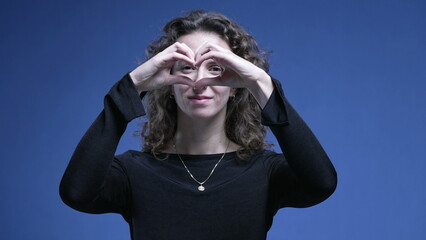 Woman doing heart shape with hands standing on blue background looking directly at camera. 20s...