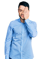 Hispanic man with beard wearing casual business shirt covering one eye with hand, confident smile on face and surprise emotion.