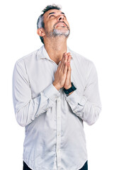 Middle age hispanic with grey hair wearing casual white shirt begging and praying with hands together with hope expression on face very emotional and worried. begging.