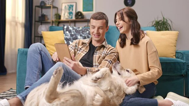 Positively Caucasian people resting together at home. Attractive husband using tablet device while pretty woman petting funny labrador while laying on floor. Lovely couple taking shared family photo.