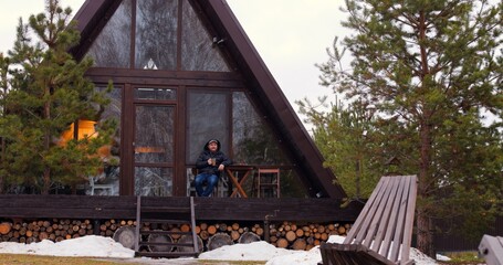 Man enjoying hot beverage on terrace of wooden house in forest, epitome of relaxation. Serene setting for unwinding, perfect for city escape and relaxation. Savor moments of relaxation in nature lap.