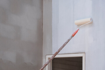 Long handle roller brush applying primer white paint with door frame on cement wall inside of house construction site, building and home renovation concept
