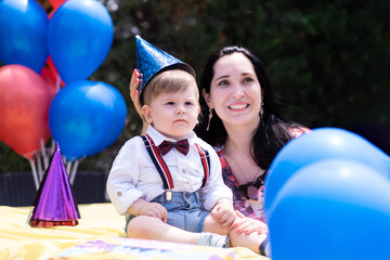 Fototapeta na wymiar Mother son first year birthday, toddler style with suspenders, first birthday, balloons, party hats, and an assortment of birthday decorations. 