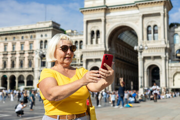 Beautiful senior blonde woman taking selfie on Duomo square, Galleria Vittorio Emanuele II shopping arcade in Milan, Italy. Traveling Europe in summer. Attractive 60s female is exploring city