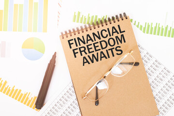 In a craft colour notebook is a FINANCIAL FREEDOM AWAITS inscription, next to pencils, glasses,...