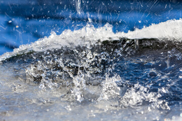 Meltwater drips onto the water surface in a rain barrel rimmed with ice crystals. Vivid water surface with drops and splashes in bright contrasting sunlight. Close up with selective focus and motion.