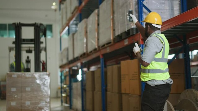 African American male worker going through inventory list while checking stock at distribution warehouse.Retail Warehouse full of Shelves with Goods in Cardboard Boxes.African male forklift operator