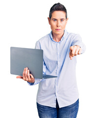 Young woman with short hair holding laptop pointing with finger to the camera and to you, confident gesture looking serious