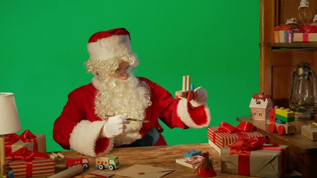 Santa Claus at the table with gifts and paints a wooden toy on a green background.