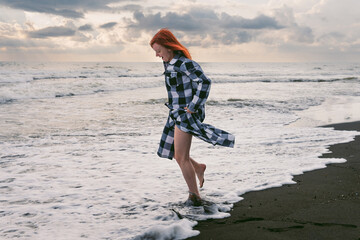 A young woman in a long checkered shirt stands on the black sand. feet in cold water, stormy waves of the sea. Heavy gray rain clouds at sunset. - Powered by Adobe